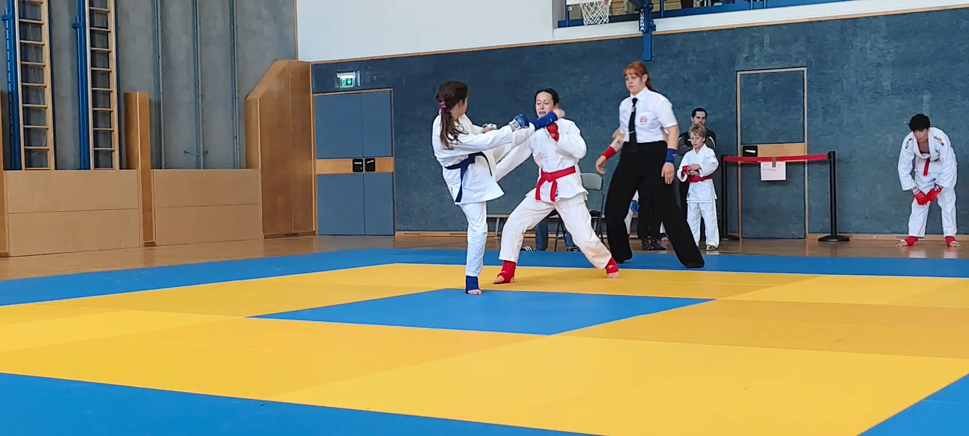 Zwei Kämpferinnen im Jiu-Jitsu-Wettkampf auf blau-gelber Matte, daneben die Kampfrichterin